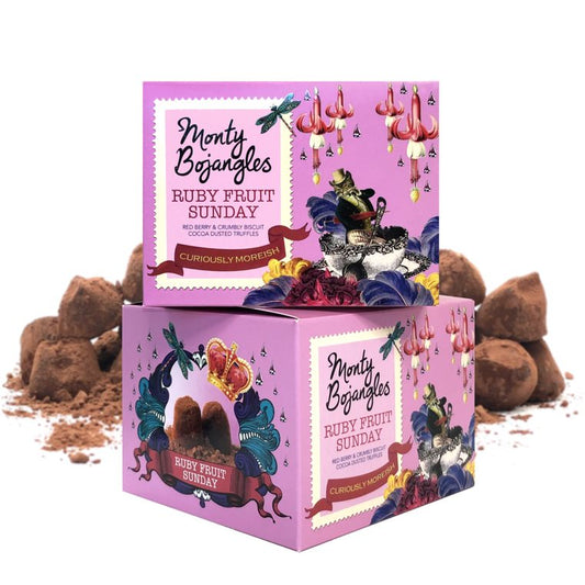 Op de foto staan twee doosjes van de Monty Bojangles in de smaak Ruby Fruit Sunday. De doosjes zijn lichtroze. Achter de doosjes liggen wat chocoladetruffels.