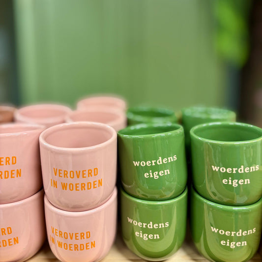 Op de foto staan links lichtroze mokken met 'Veroverd in Woerden'. Rechts staan groene mokken met de tekst 'Woerdens eigen'.