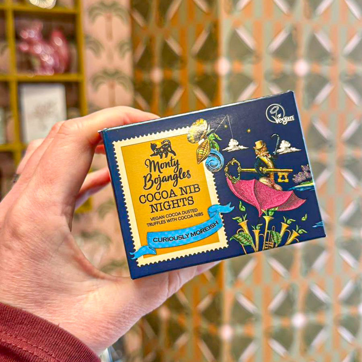 Een donkerblauw doosje met Monty Bojangles chocoladetruffels in de smaak Cocoa Nib Nights. Het doosje wordt vastgehouden door een hand.