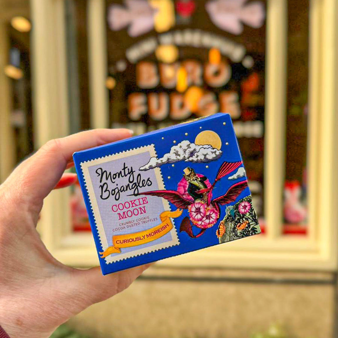 Een hand houdt een kobaltblauw doosje vast met Monty Bojangles chocoladetruffels in de smaak Cookie Moon.