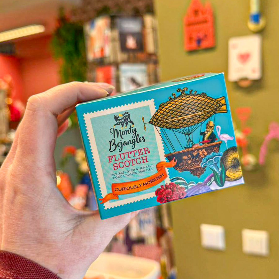Een hand houdt een doosje met Monty Bojangles Flutter Scotch chocoladetruffels vast. Het doosje is lichtblauw met oranje details. 