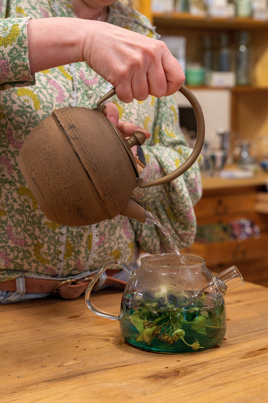 Op de foto zie je een hand met een bruine theepot die water in een glazen theepot schenkt. In de glazen theepot kleurt het water blauw-groen door de kruiden die erin zitten.
