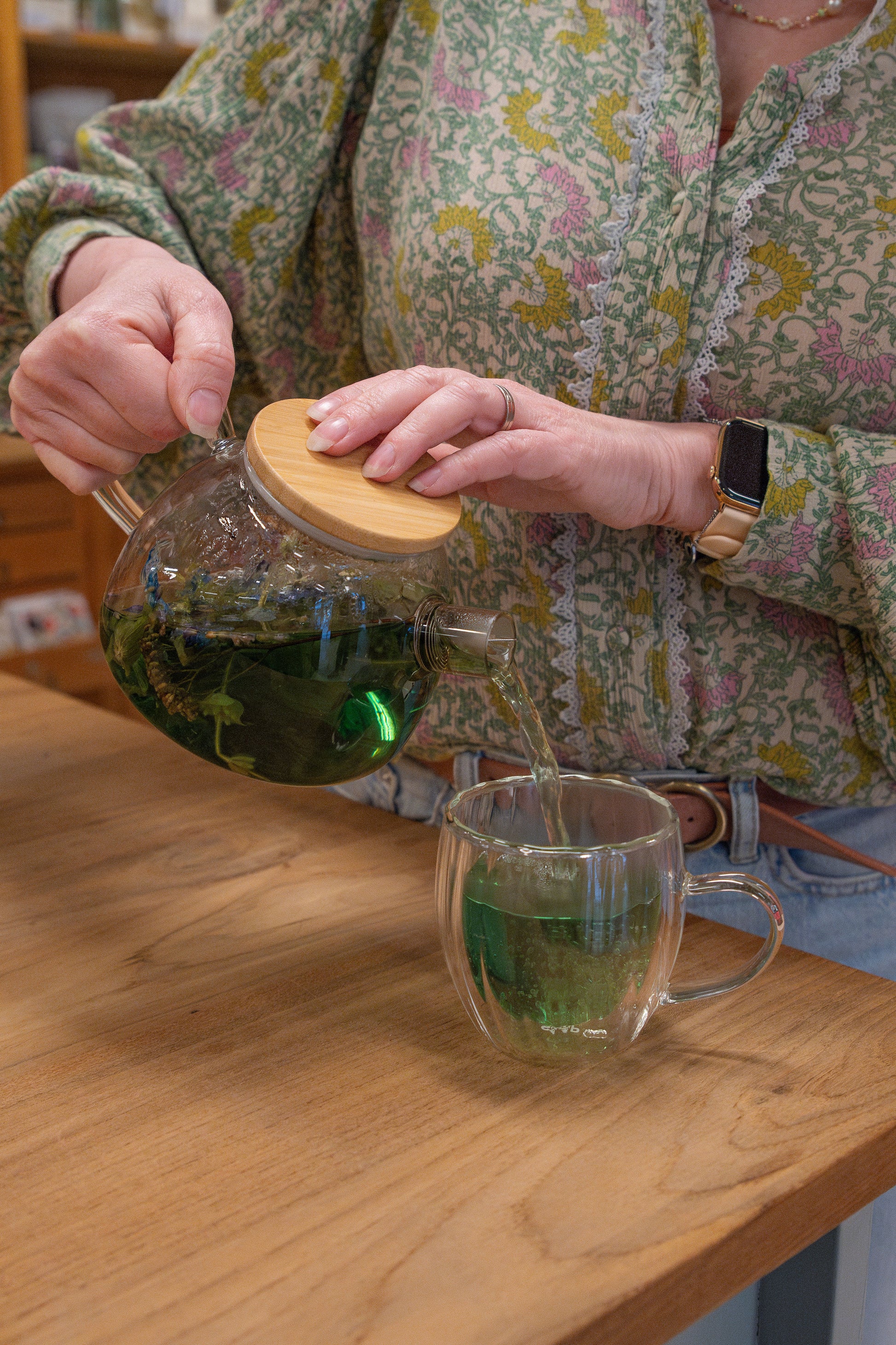 Op de foto staan twee handen die een groene kruidenthee vanuit een glazen theepot in een theeglas schenken. 