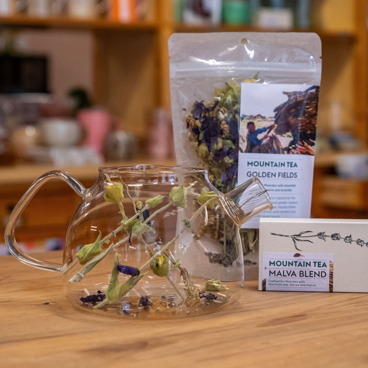 Op de foto staat een theepot gevuld met kruiden. Daarbij staat een klein doosje en een zakje met Mountain Tea.