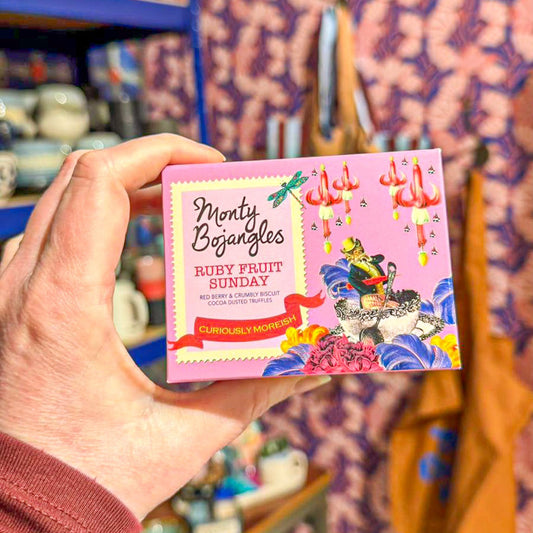 Op de foto staat een hand die een doosje Monty Bojangles in de smaak Ruby Fruit Sunday vasthoudt. De Monty Bojangles zitten in een lichtroze doosje met allerlei illustraties erop. 