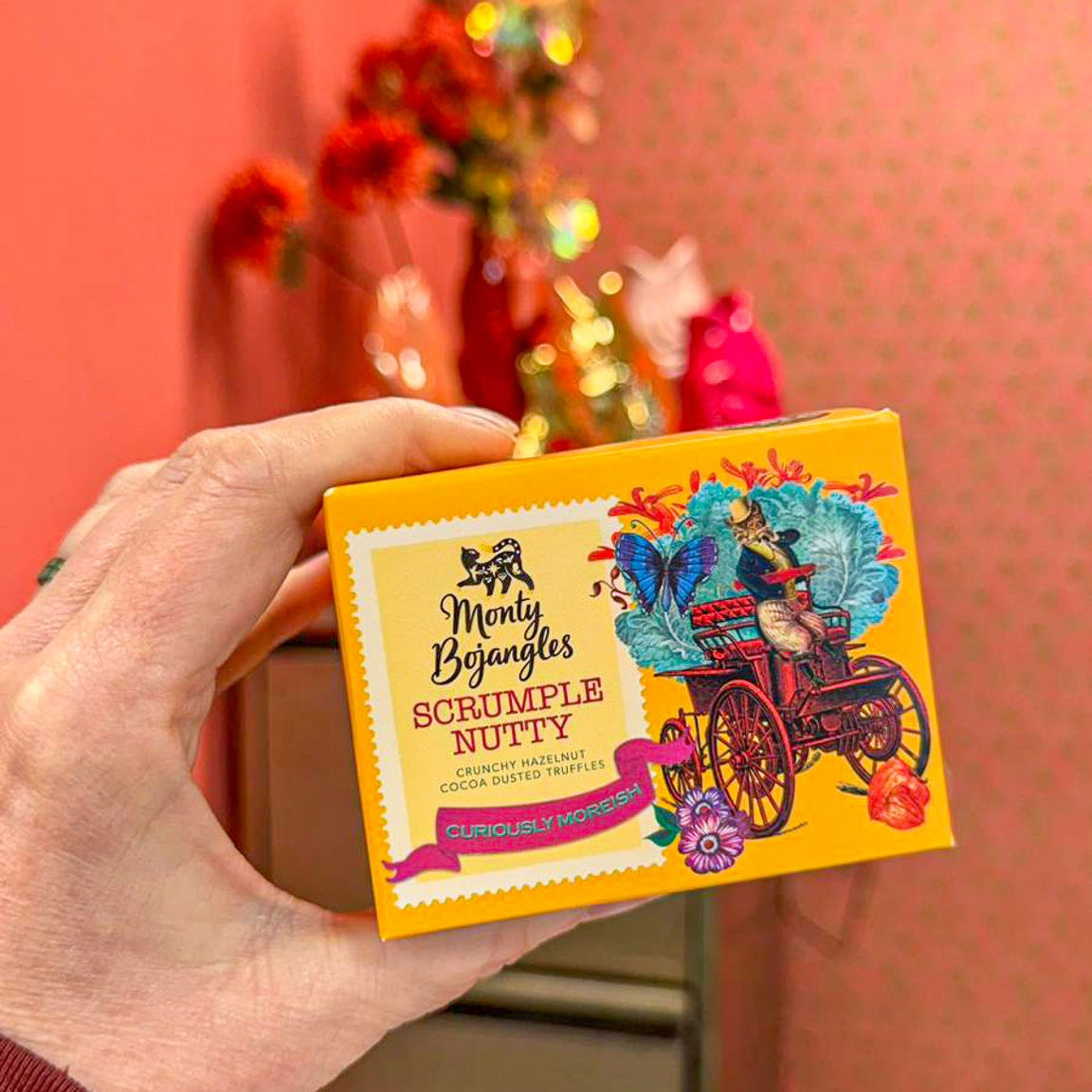 Op de foto staat een hand die een geel doosje Monty Bojangles vasthoudt in de smaak Scrumple Nutty. 