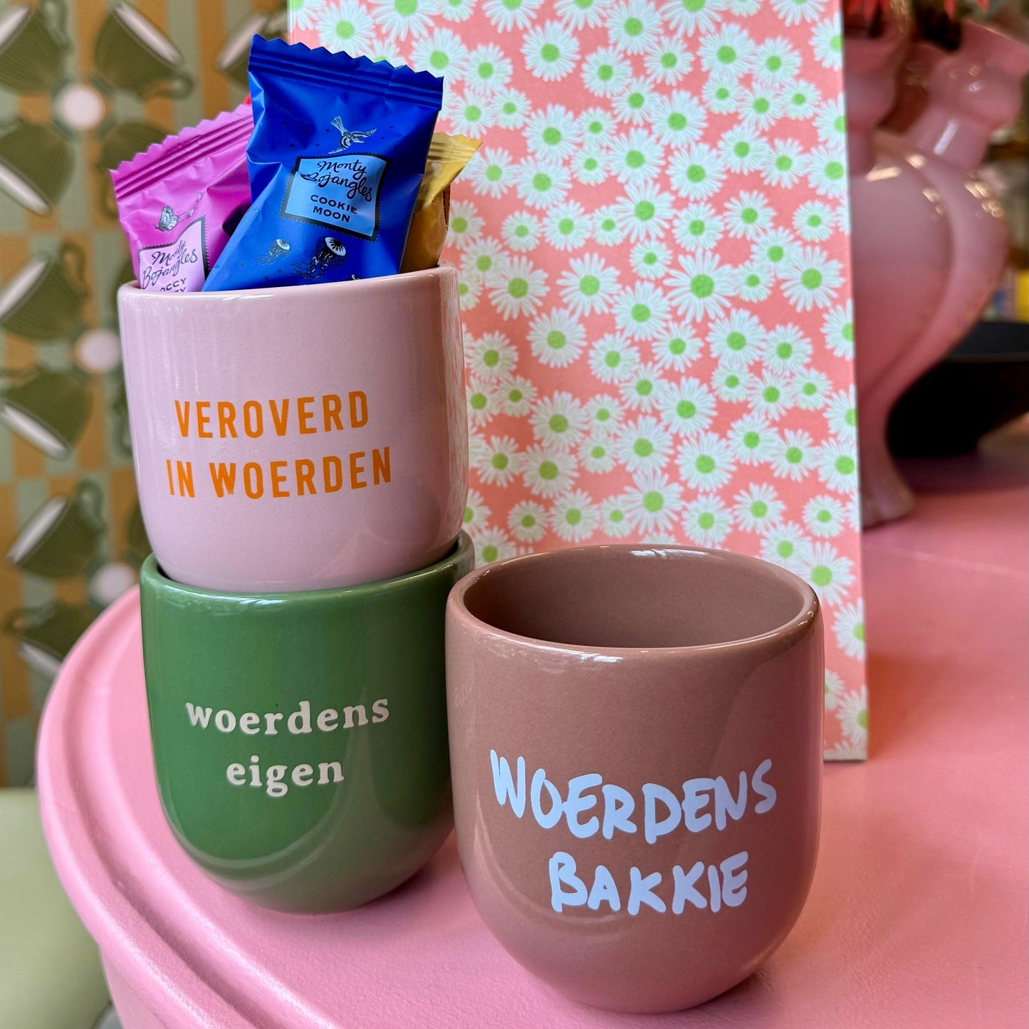 Op de foto zie je drie gekleurde mokken; een groene, roze en een taupekleurige. Op de achtergrond staat een cadeauzakje met bloemen.