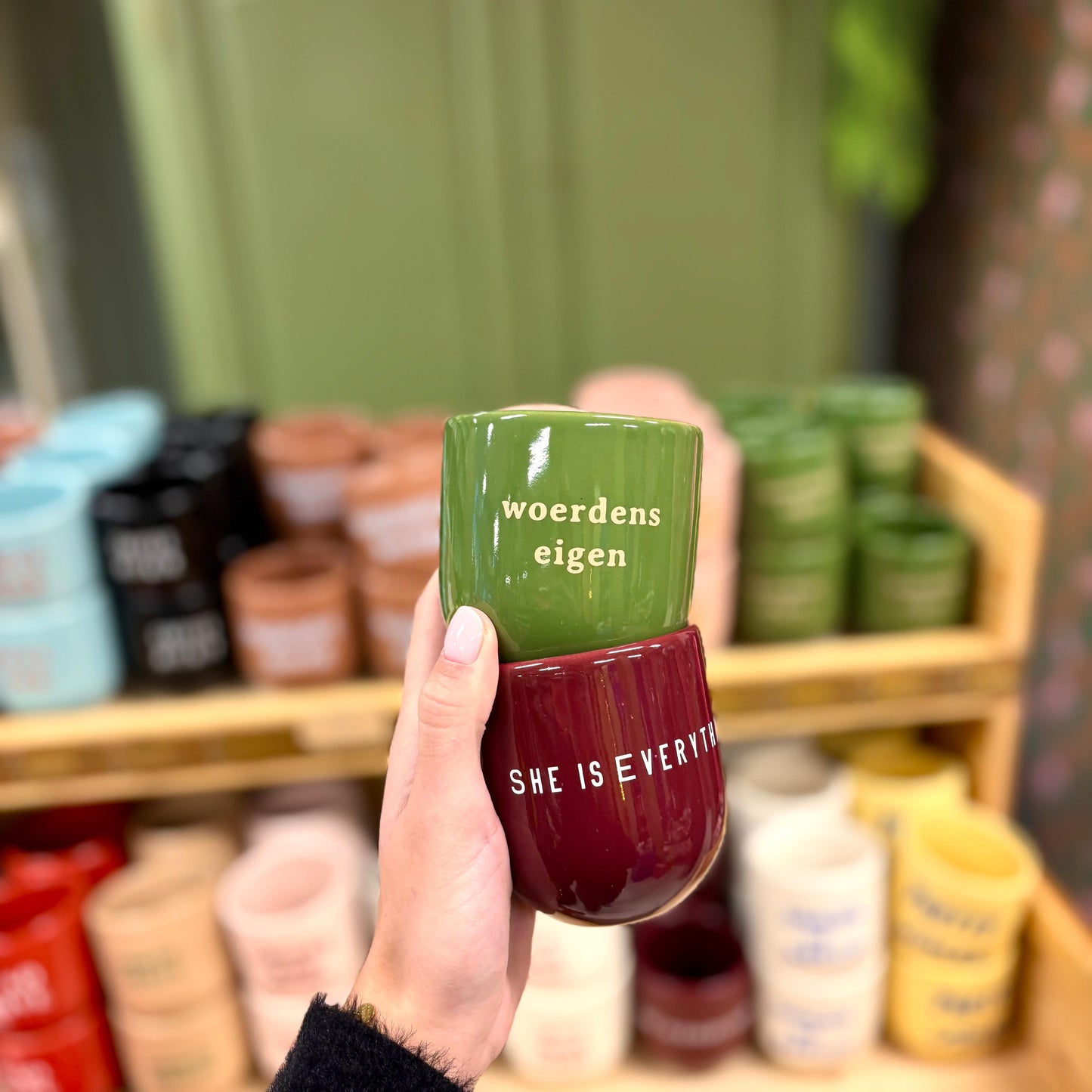 Op de foto staat een hand die twee mokken vasthoudt. Een groene mok met de tekst 'Woerdens eigen', op de donkerrode mok staat 'She is everything'.